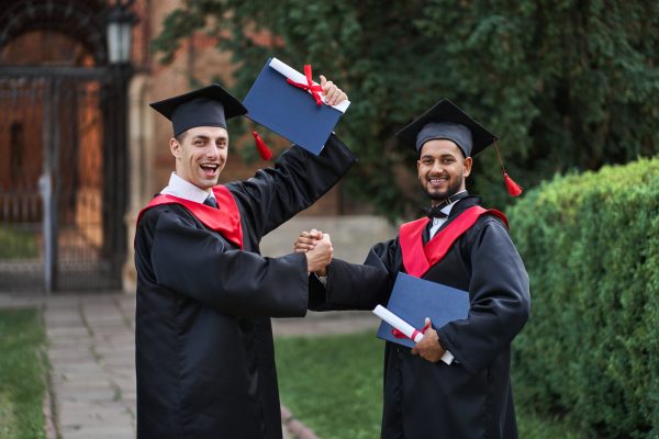 Two international graduate celebrating graduation in university campus and looking at camera.